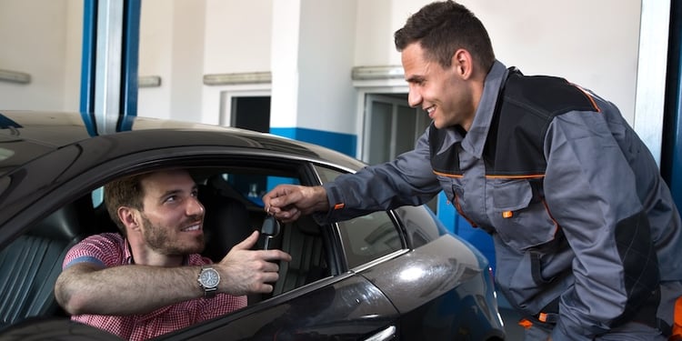 A male automotive technician hands the keys to a happy customer sitting in their vehicle after service.